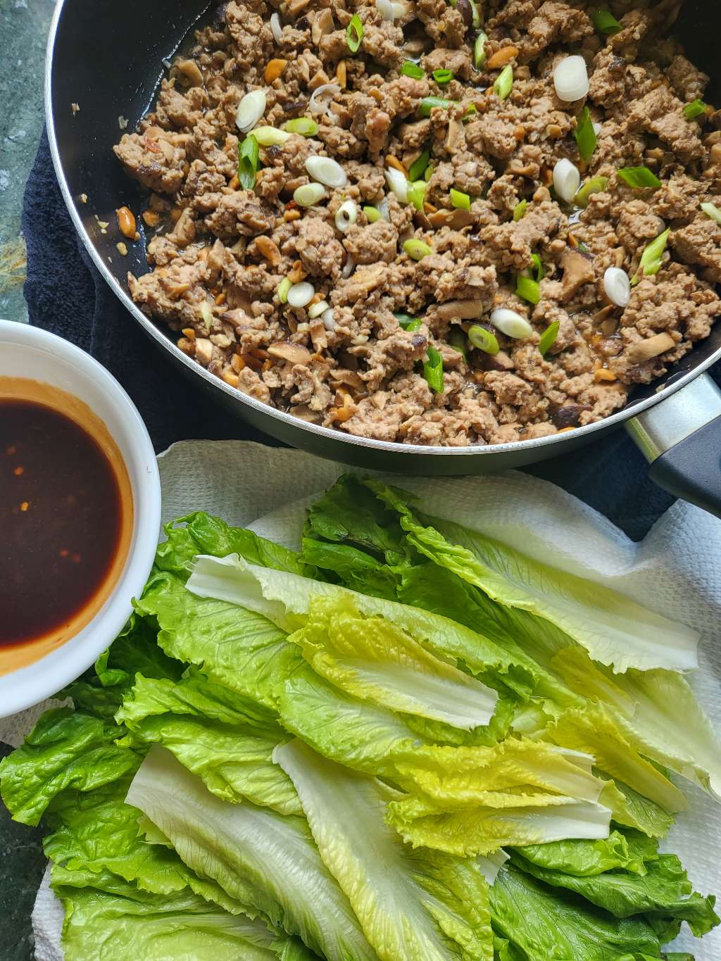 Another photo of the cooked ground pork mixture in a pan, next to some lettuce leaves on a place, and dipping sauce in a bowl