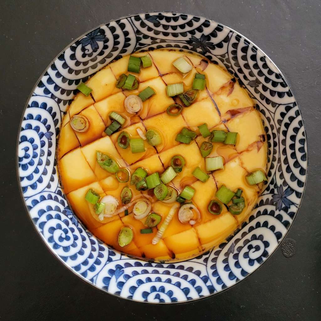 Overhead photo of steamed eggs with slices in them and topped with scallions and soy sauce