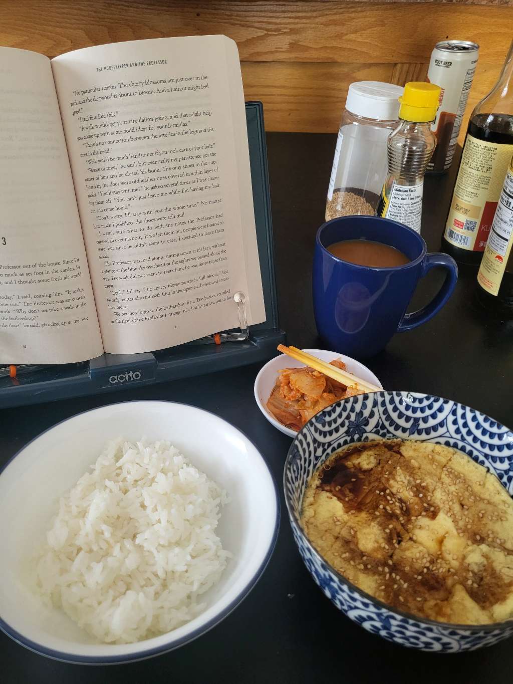 Some microwaved steamed eggs in a bowl, topped with soy sauce and sesame seeds. There is also rice, kimchi, and a book in the back