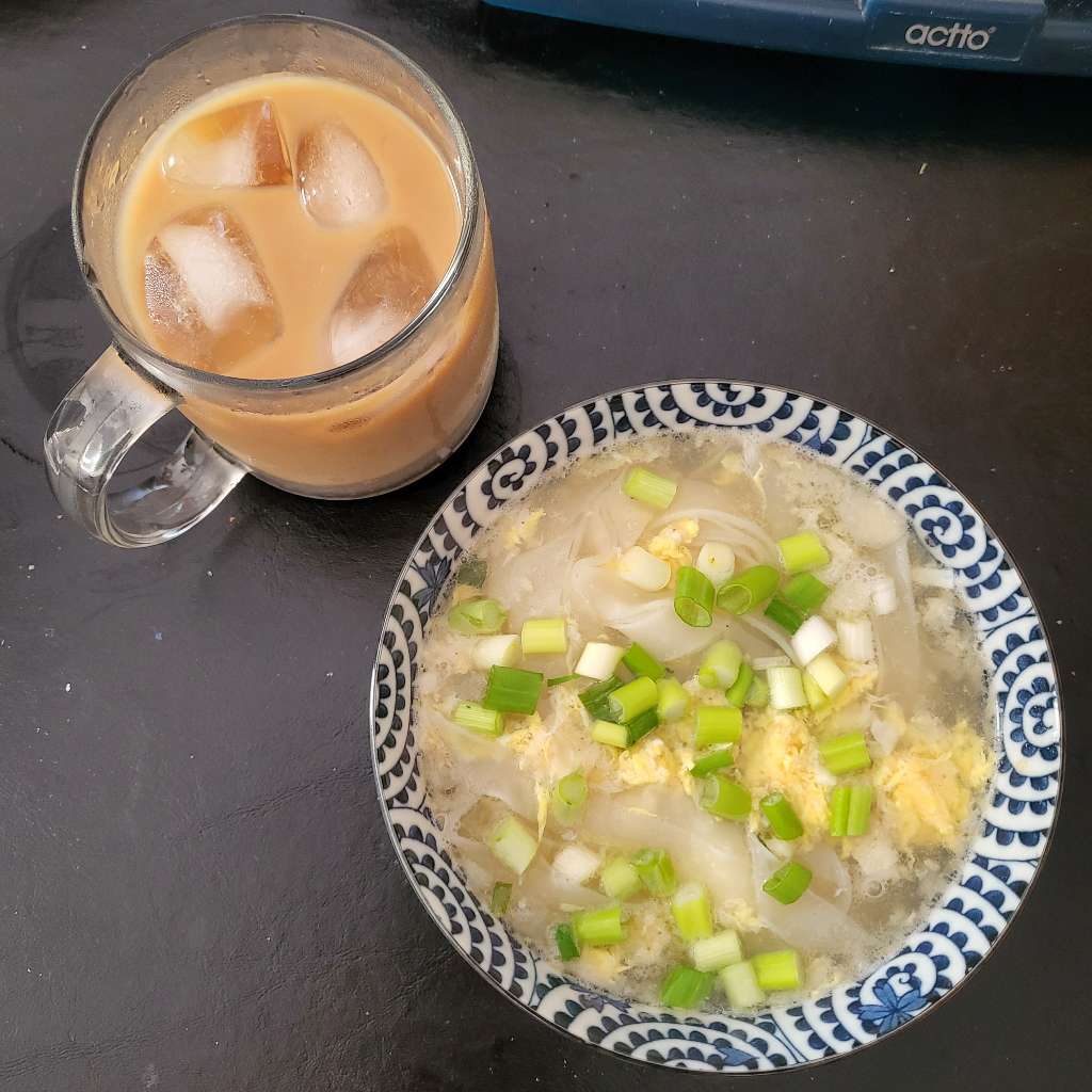 An overhead shot of the bowl of noodles, topped with scallions, along with a cup of iced coffee next to it